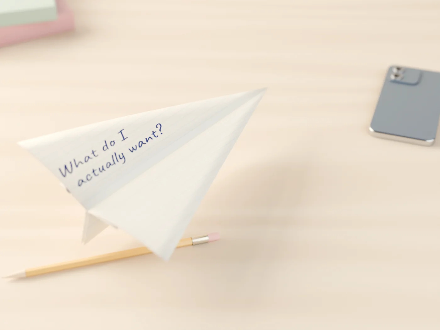 A paper plane folded from a notebook page that asks “What do I actually want?”, on a desk beside a phone turned face-down